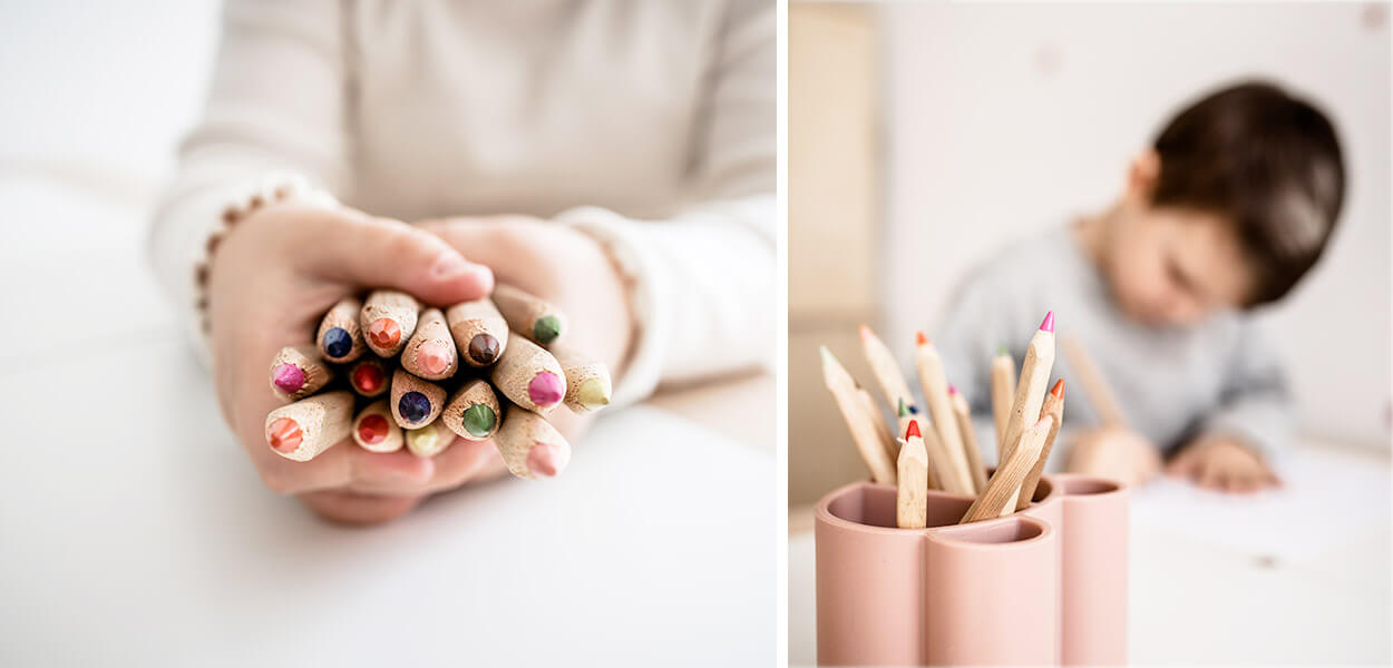 Kind hält viele Buntstifte, Buntstifte in Stiftebox und malender Junge im Hintergrund